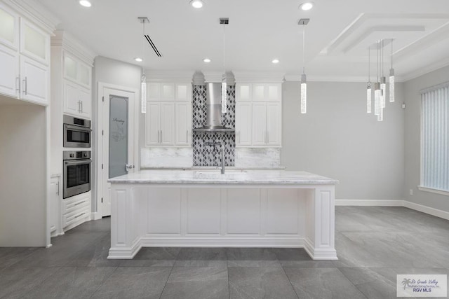 kitchen featuring white cabinets, an island with sink, hanging light fixtures, and wall chimney range hood