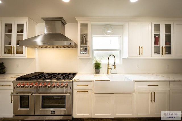 kitchen featuring wall chimney exhaust hood, sink, white cabinetry, light stone countertops, and range with two ovens