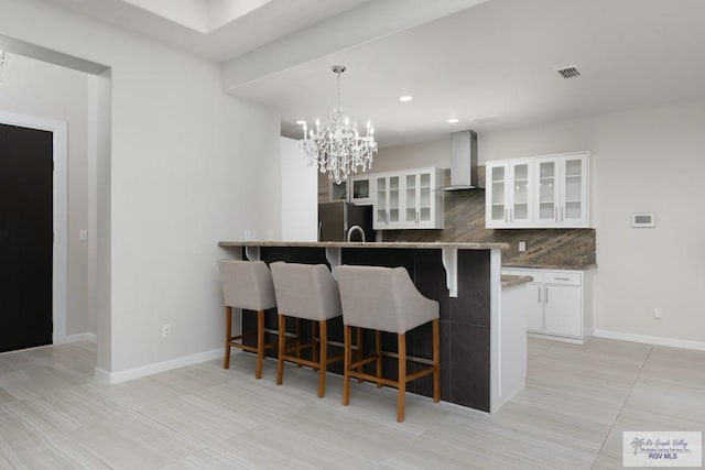 kitchen featuring wall chimney range hood, stainless steel fridge, white cabinetry, backsplash, and hanging light fixtures