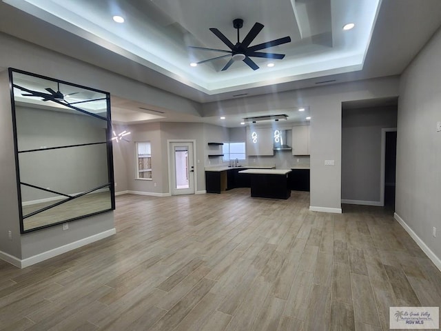 unfurnished living room with ceiling fan, sink, a tray ceiling, and light hardwood / wood-style floors