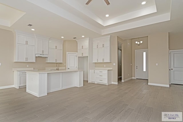 kitchen with light wood-type flooring, ceiling fan with notable chandelier, a raised ceiling, a kitchen island with sink, and white cabinets