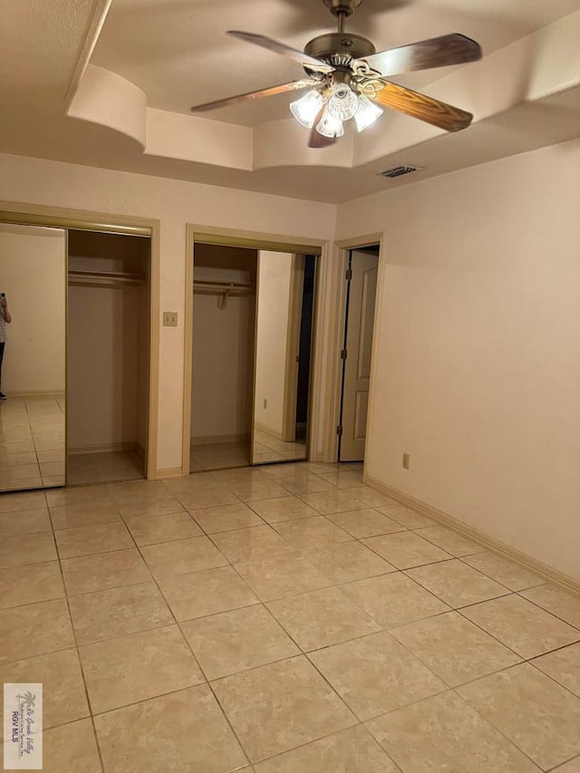 unfurnished bedroom with a tray ceiling, ceiling fan, light tile patterned floors, and two closets