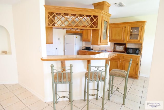 9165 Los Olmos Dr, Los Fresnos, TX 78566 - kitchen featuring freestanding refrigerator, black microwave, a kitchen bar, glass insert cabinets, and a peninsula