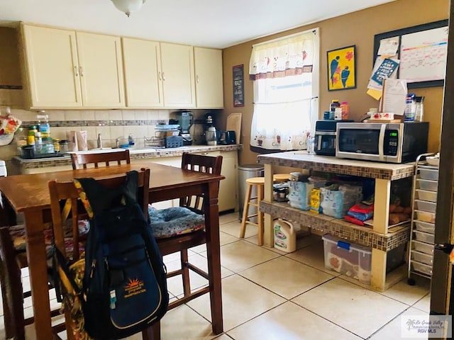 kitchen with light tile patterned floors, decorative backsplash, stainless steel microwave, light countertops, and cream cabinetry