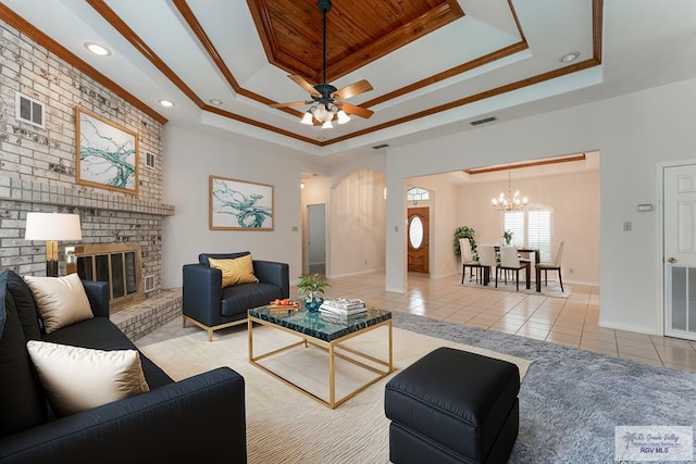 tiled living room featuring a brick fireplace, a tray ceiling, ceiling fan with notable chandelier, and ornamental molding