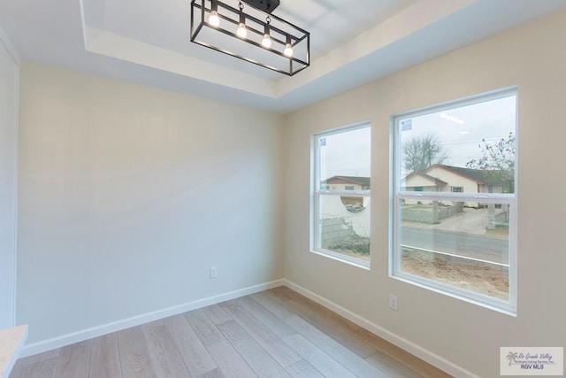 empty room with baseboards, light wood-type flooring, a raised ceiling, and a notable chandelier