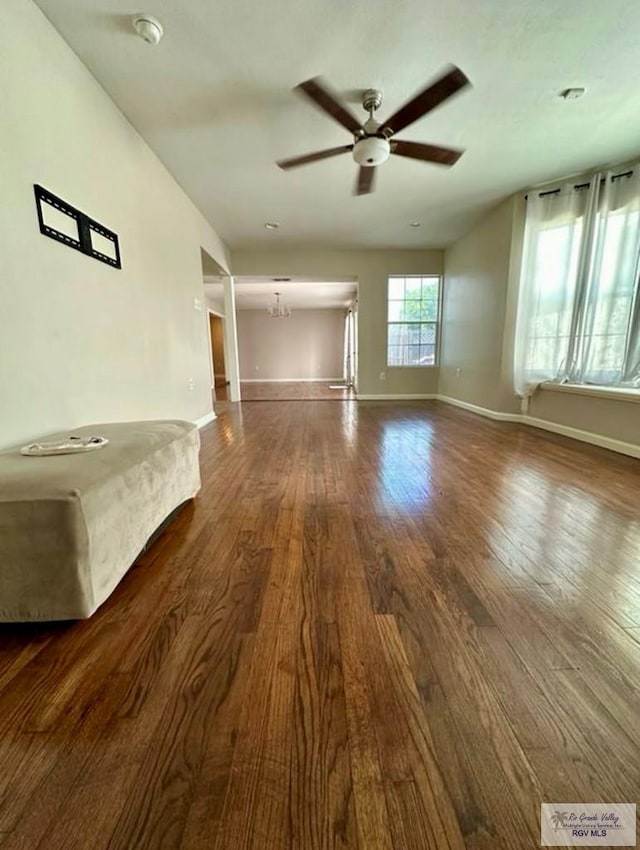 unfurnished living room with ceiling fan with notable chandelier, baseboards, and dark wood-style flooring