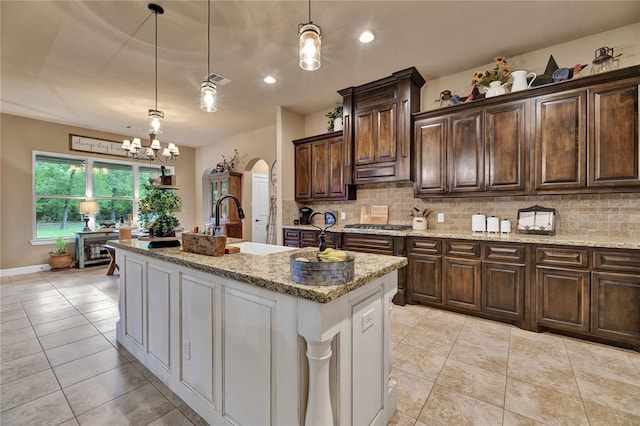 kitchen featuring light stone counters, backsplash, a notable chandelier, decorative light fixtures, and a center island with sink