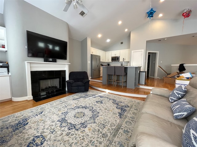 living room with a tiled fireplace, ceiling fan, light wood-type flooring, and high vaulted ceiling