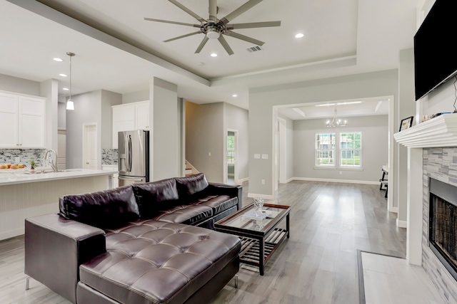 living room featuring light hardwood / wood-style flooring, a fireplace, ceiling fan with notable chandelier, sink, and a raised ceiling