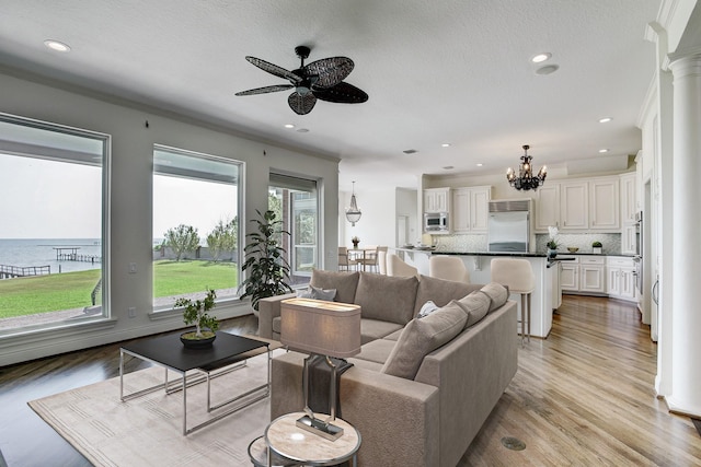 living room featuring ceiling fan with notable chandelier, a water view, light hardwood / wood-style flooring, and ornate columns