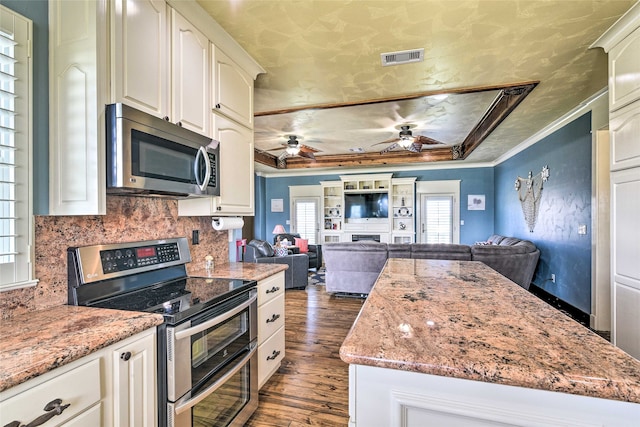 kitchen with dark wood-type flooring, stainless steel appliances, ceiling fan, ornamental molding, and a tray ceiling