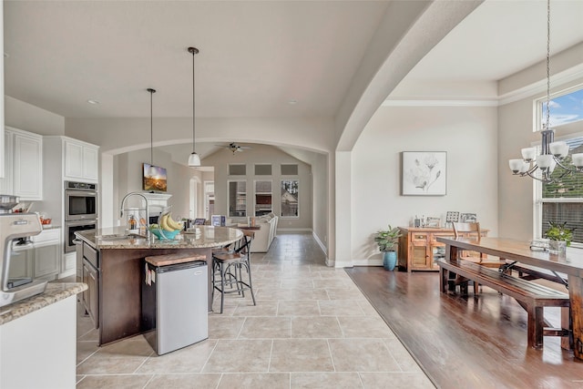 kitchen featuring white cabinetry, hanging light fixtures, ceiling fan with notable chandelier, a kitchen island with sink, and light tile floors