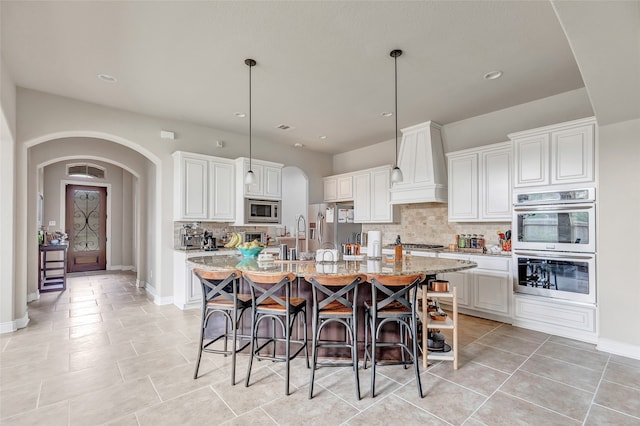 kitchen with stainless steel appliances, custom exhaust hood, a center island with sink, backsplash, and hanging light fixtures