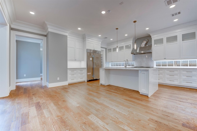 kitchen with built in refrigerator, a center island with sink, pendant lighting, wall chimney range hood, and white cabinets