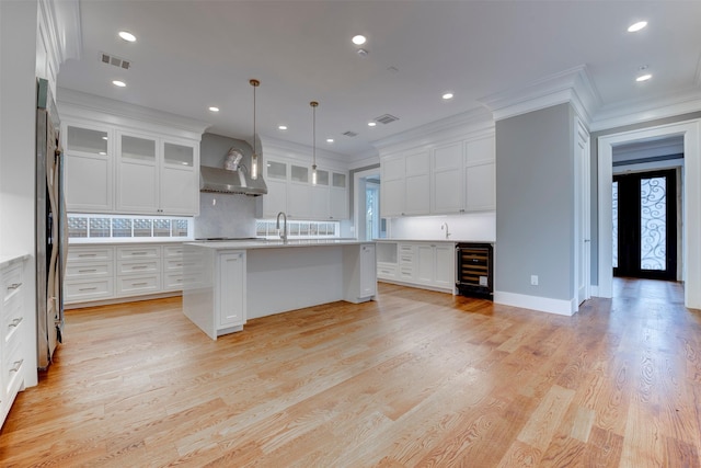 kitchen with pendant lighting, white cabinets, beverage cooler, a kitchen island with sink, and wall chimney range hood