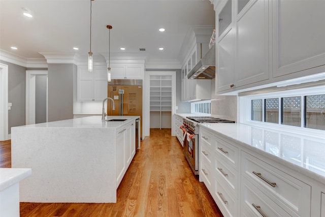 kitchen featuring wall chimney exhaust hood, white cabinetry, decorative light fixtures, a center island with sink, and premium appliances