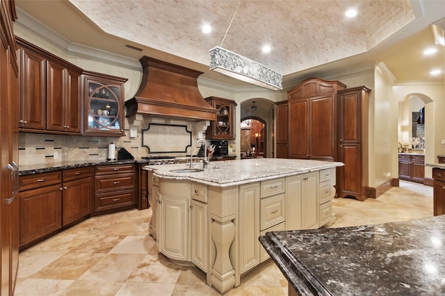 kitchen with custom exhaust hood, backsplash, a kitchen island with sink, cream cabinets, and dark stone countertops