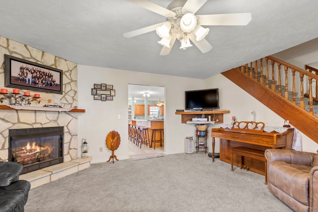 living room with a textured ceiling, light colored carpet, a stone fireplace, and ceiling fan