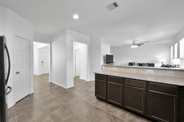 kitchen with stainless steel fridge, ceiling fan, light tile flooring, light stone countertops, and dark brown cabinetry