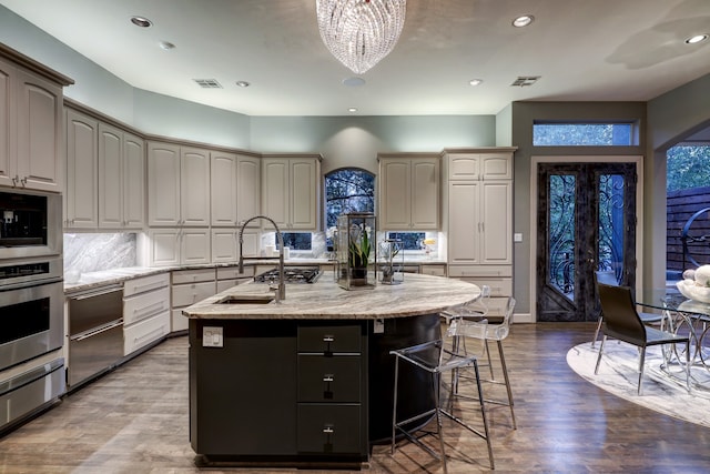 kitchen with an inviting chandelier, hardwood / wood-style flooring, an island with sink, light stone countertops, and backsplash