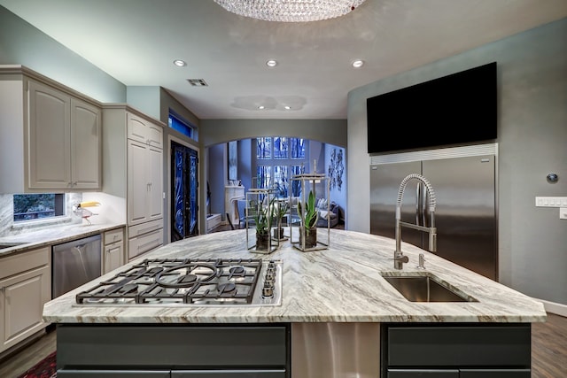 kitchen featuring an inviting chandelier, dark wood-type flooring, appliances with stainless steel finishes, light stone counters, and a center island with sink