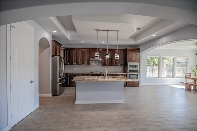 kitchen with a center island with sink, appliances with stainless steel finishes, a tray ceiling, and light stone countertops