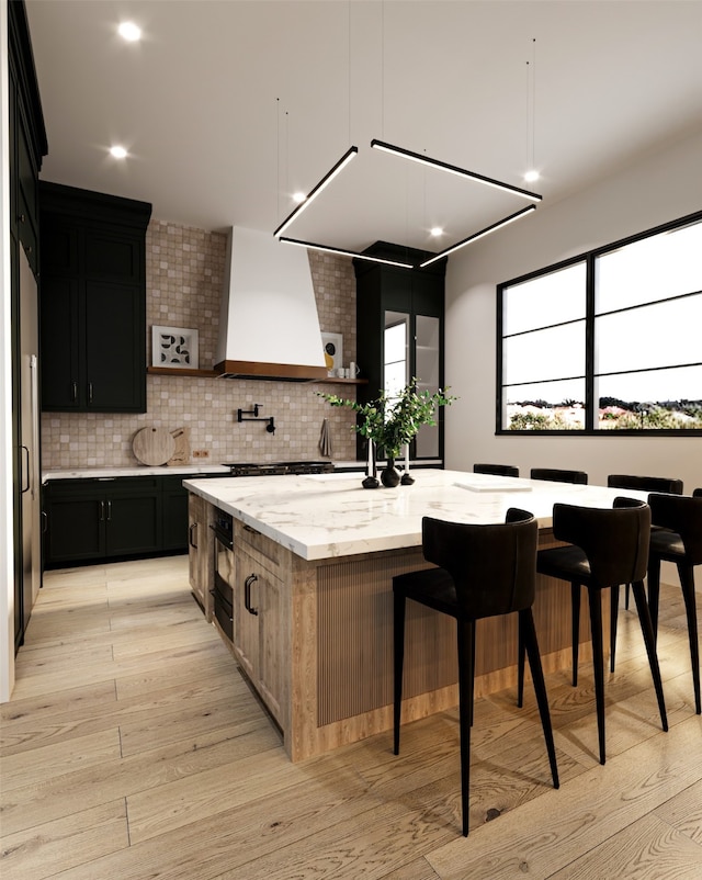 kitchen with light stone counters, light hardwood / wood-style floors, tasteful backsplash, custom exhaust hood, and a center island