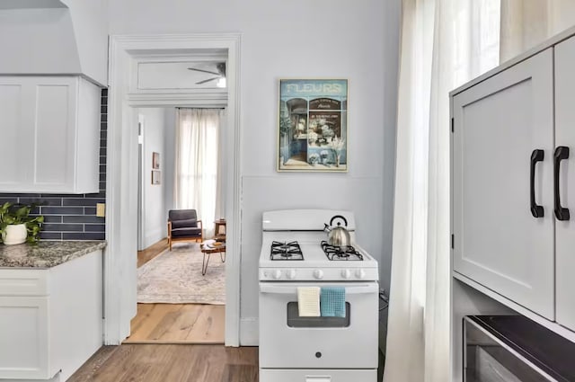 kitchen with backsplash, white gas stove, dark stone counters, wood finished floors, and white cabinetry