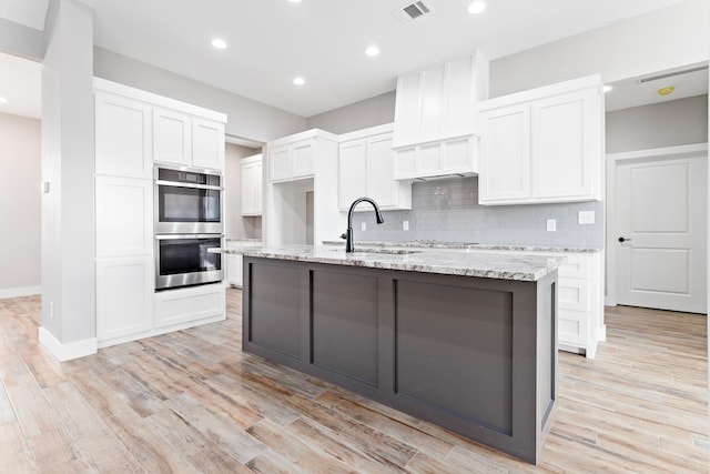 kitchen featuring light hardwood / wood-style floors, stainless steel double oven, white cabinets, backsplash, and a kitchen island with sink