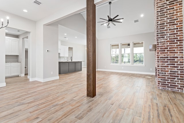 unfurnished living room with lofted ceiling, brick wall, light wood-type flooring, and ceiling fan with notable chandelier