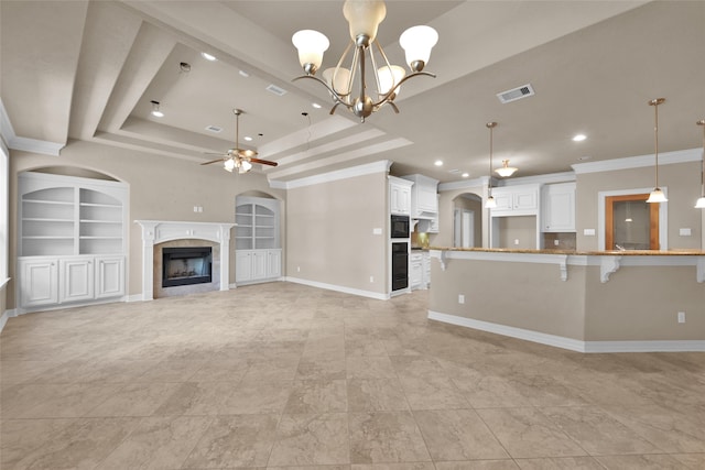 unfurnished living room with a raised ceiling, ceiling fan with notable chandelier, built in shelves, a fireplace, and crown molding