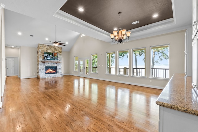 unfurnished living room featuring a stone fireplace, a tray ceiling, light hardwood / wood-style floors, and ceiling fan with notable chandelier