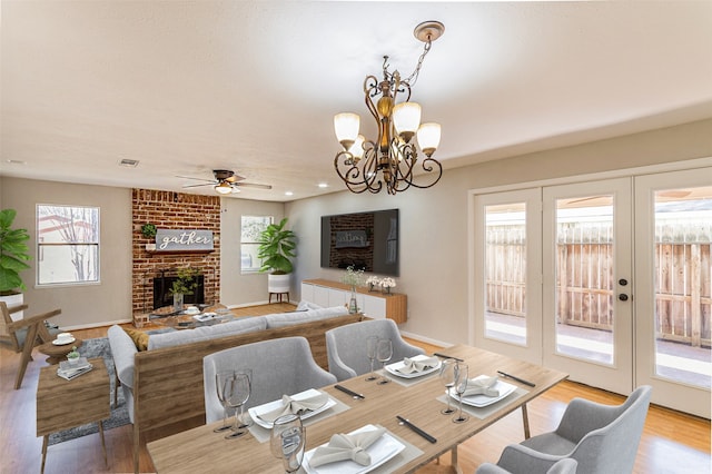 dining area featuring ceiling fan with notable chandelier, light wood-type flooring, brick wall, french doors, and a brick fireplace