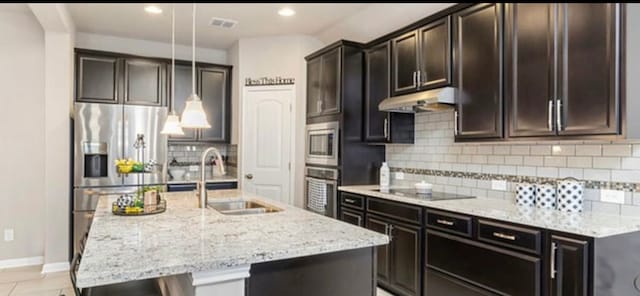 kitchen featuring light stone countertops, backsplash, hanging light fixtures, stainless steel appliances, and a kitchen island with sink