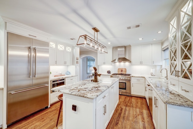 kitchen featuring pendant lighting, high end appliances, wall chimney exhaust hood, a kitchen island, and white cabinetry