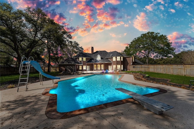 pool at dusk with a diving board, a water slide, and a patio area