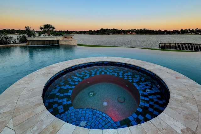 pool at dusk with a patio area, an in ground hot tub, pool water feature, and a water view