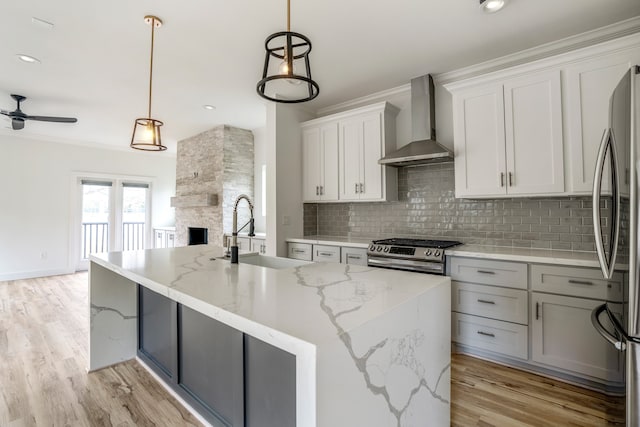 kitchen with ceiling fan, appliances with stainless steel finishes, light hardwood / wood-style flooring, a stone fireplace, and wall chimney exhaust hood