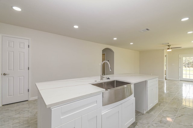 kitchen with ceiling fan, white cabinets, light stone counters, sink, and a center island with sink