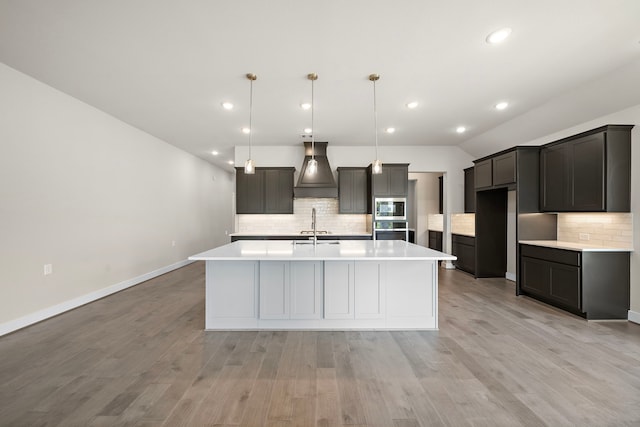 kitchen featuring premium range hood, an island with sink, decorative light fixtures, stainless steel appliances, and light wood-type flooring