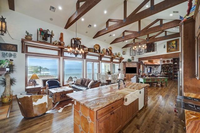 kitchen with dark hardwood / wood-style floors, beamed ceiling, a kitchen island with sink, a notable chandelier, and light stone counters