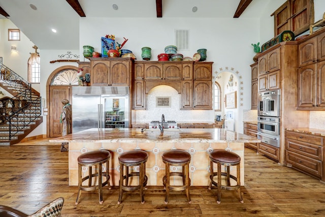 kitchen with an island with sink, stainless steel appliances, tasteful backsplash, wood-type flooring, and beamed ceiling