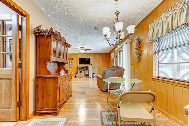 dining space featuring ceiling fan with notable chandelier, a textured ceiling, light hardwood / wood-style flooring, and wood walls