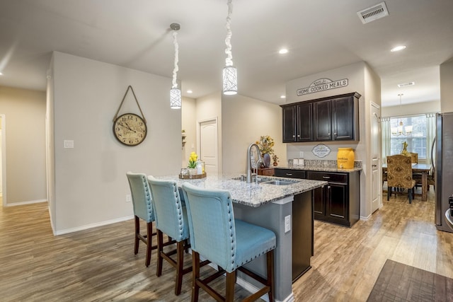 kitchen with light wood-type flooring, a kitchen island with sink, sink, stainless steel refrigerator, and hanging light fixtures