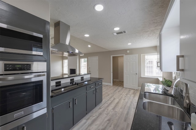 kitchen with wall chimney range hood, light hardwood / wood-style flooring, backsplash, appliances with stainless steel finishes, and sink