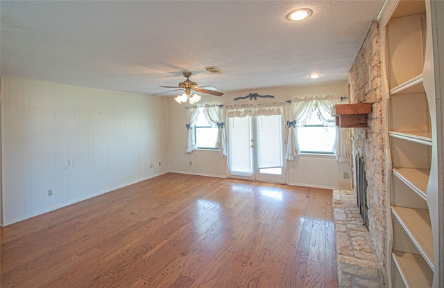 empty room with ceiling fan, light hardwood / wood-style flooring, a healthy amount of sunlight, and a stone fireplace