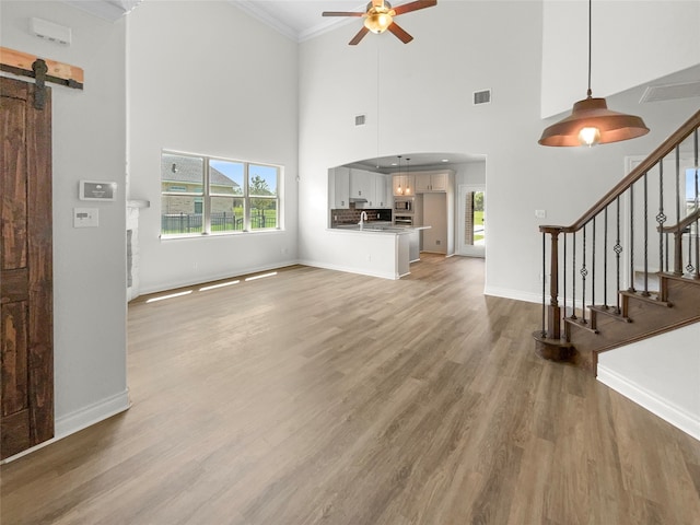 unfurnished living room with crown molding, a barn door, hardwood / wood-style floors, ceiling fan, and a towering ceiling