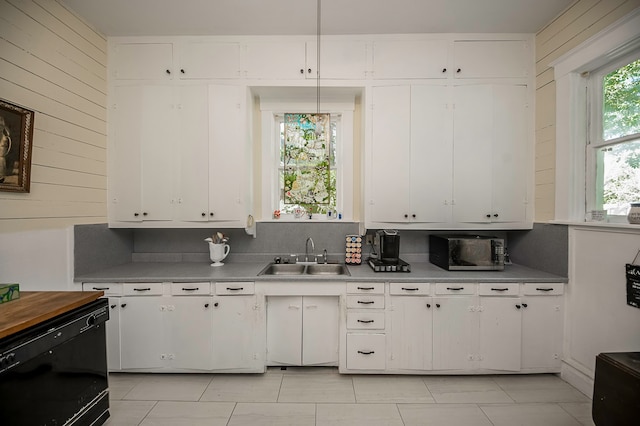 kitchen with light tile patterned flooring, white cabinets, black dishwasher, wood walls, and sink