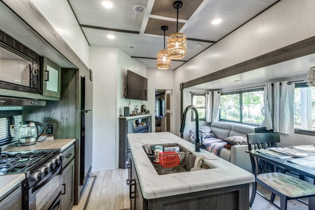 kitchen featuring a kitchen island with sink, light stone counters, black appliances, decorative light fixtures, and light wood-type flooring